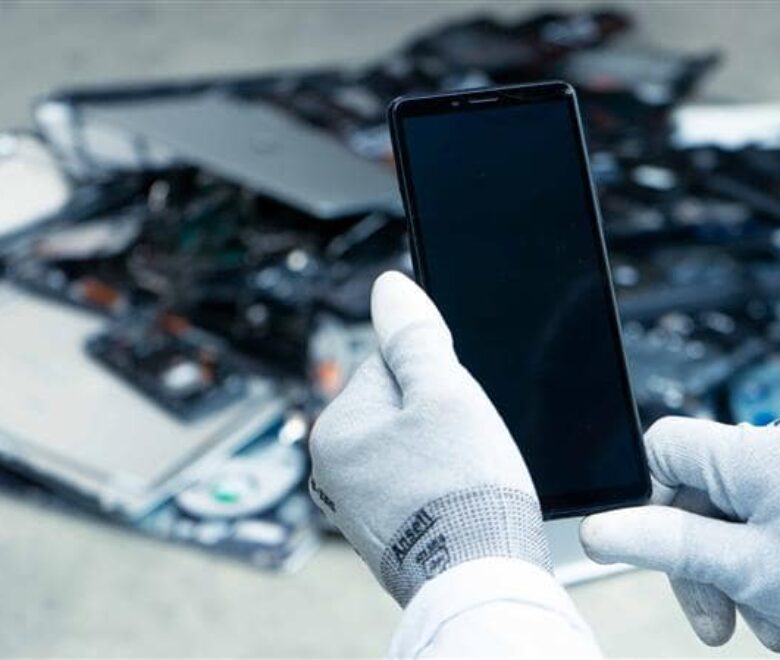 Un smartphone reconditionné tenu par les mains de Laure. Au second plan des équipements électroniques rachetés ou récupérés pour être réutilisés ou recyclés.