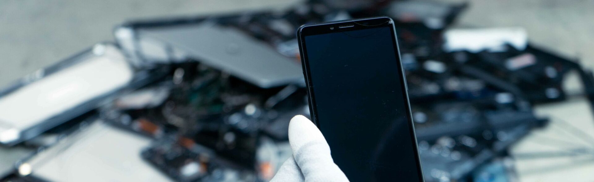 Un smartphone reconditionné tenu par les mains de Laure. Au second plan des équipements électroniques rachetés ou récupérés pour être recyclés.